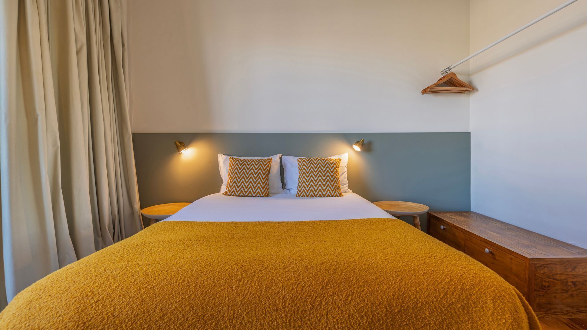 Una cama bien hecha con un edredón amarillo, dos almohadas con estampado de greca, dos mesitas de noche y una mesita auxiliar de madera.