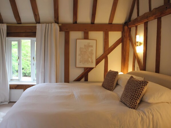 A bedroom with a large bed, wooden beams, a window with curtains, and a wall-mounted light fixture. - Headland Barn (Maldon)