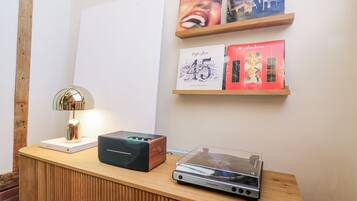 A wooden desk with a lamp, a black speaker, and a silver audio device. Above the desk, there are two framed pictures and a red book.