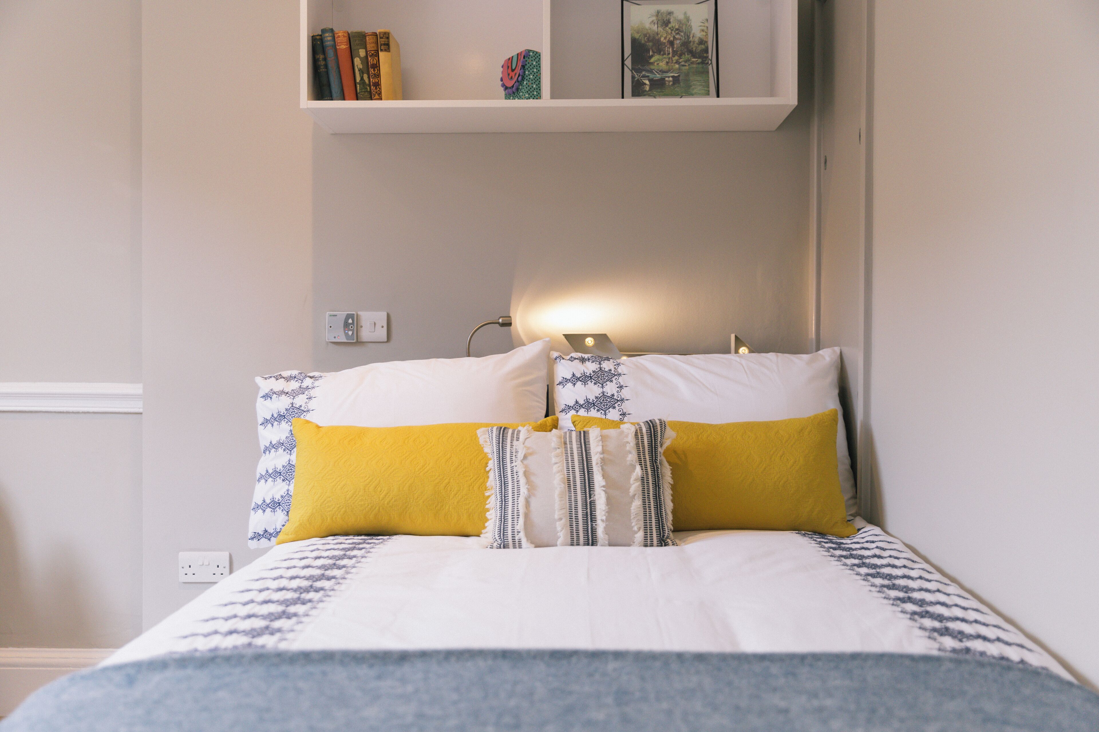 A neatly made bed with a patterned bedspread, two pillows with a yellow accent, and a bedside lamp.