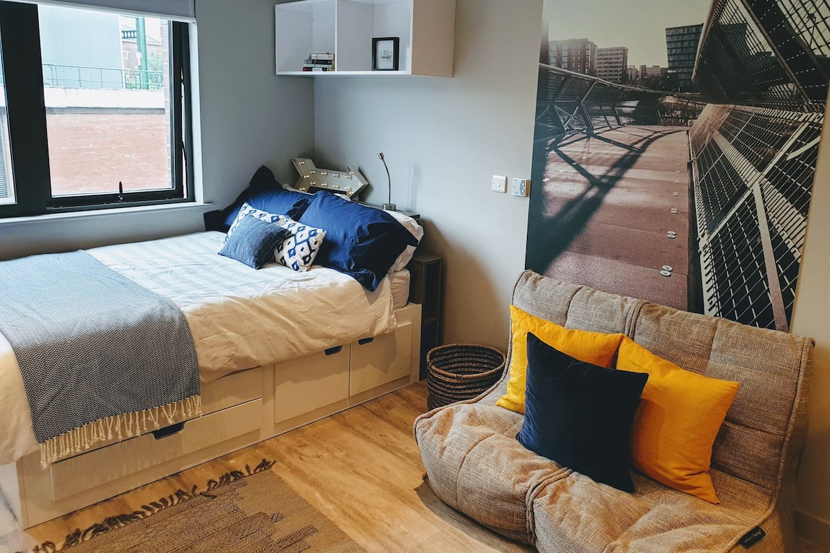 A bedroom with a bed, a sofa, a window, a wall-mounted shelf, and a cityscape mural.