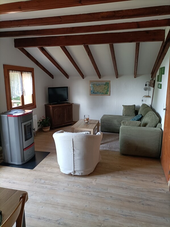 A cozy living room with exposed wooden beams, a large window with curtains, a TV, a sofa, and a fireplace. - Chalet Ida in its green setting between Gérardmer and Alsace (Anould)