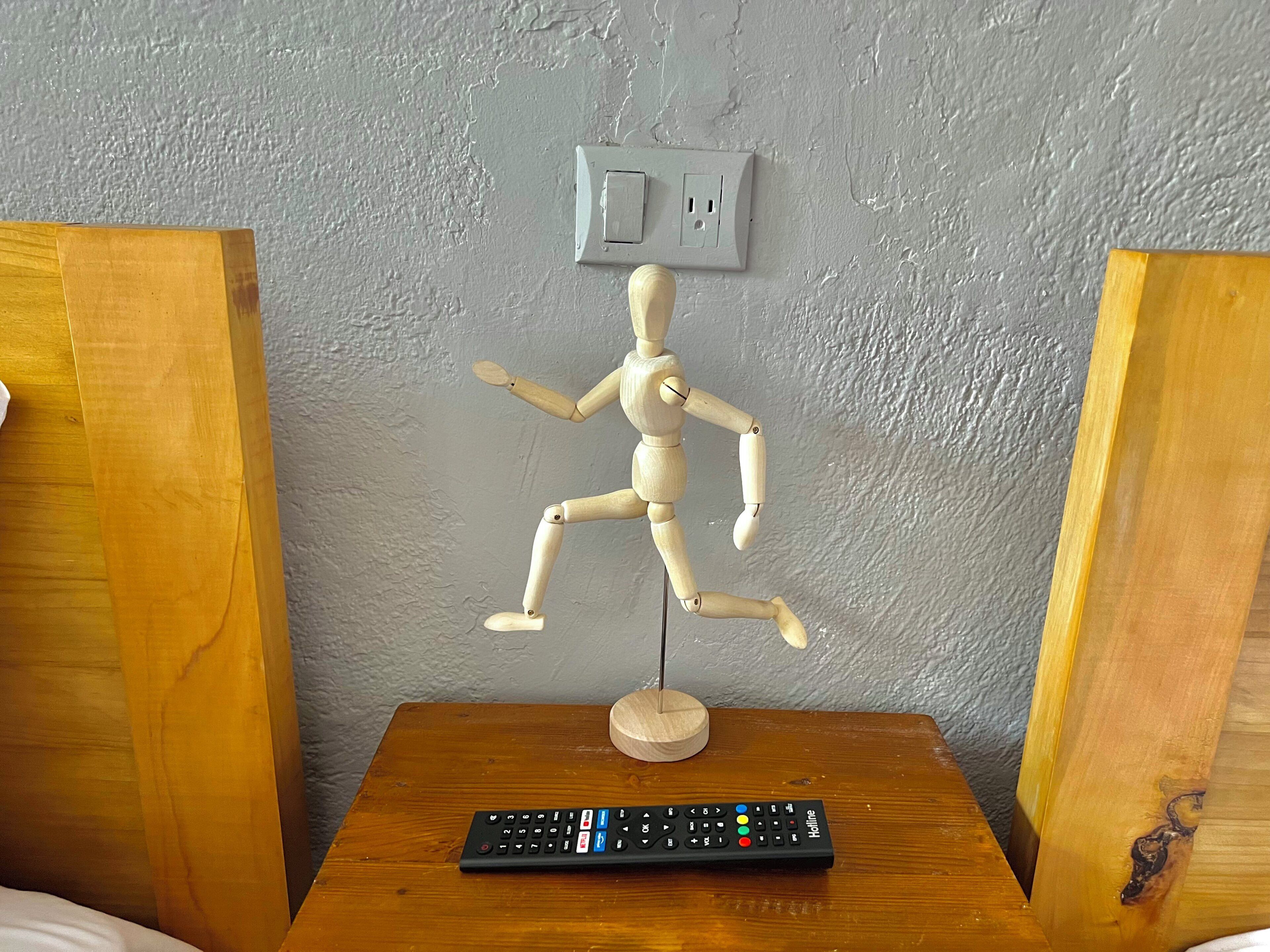 Un maniquí de madera con un control remoto sobre una mesa de madera, frente a una pared con textura.