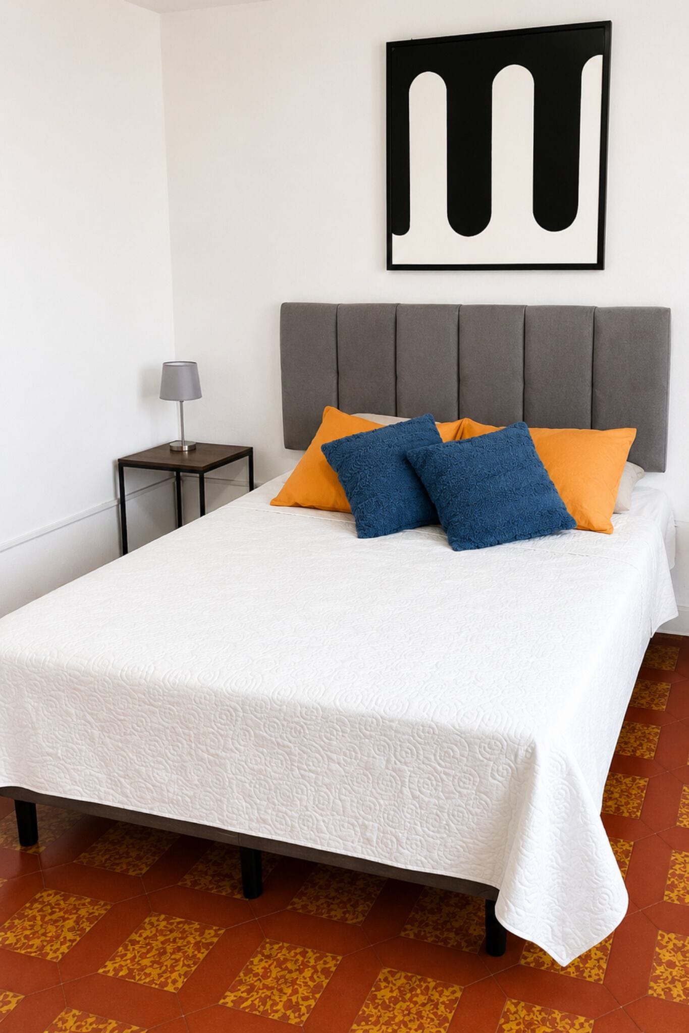 Un lit bien fait, recouvert d’une couvre-lit blanche, avec une tête de lit rembourrée de couleur grise et deux oreillers, un bleu et l’autre orange.