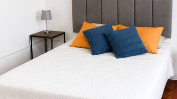 Un lit bien fait, recouvert d’une couvre-lit blanche, avec une tête de lit rembourrée de couleur grise et deux oreillers, un bleu et l’autre orange.