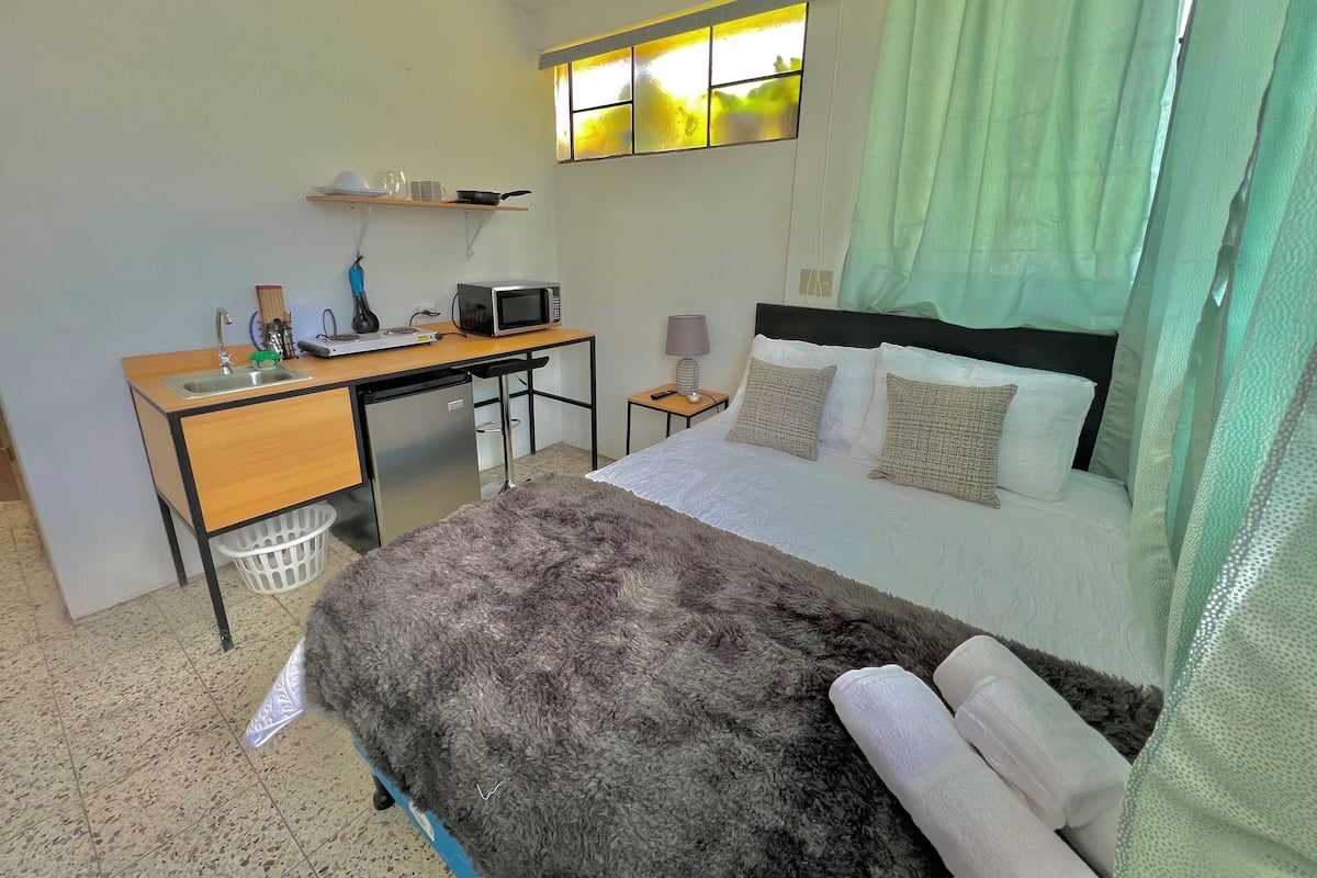 A compact hotel room with a bed, a desk with a microwave, a small table with a lamp, and a window with curtains.