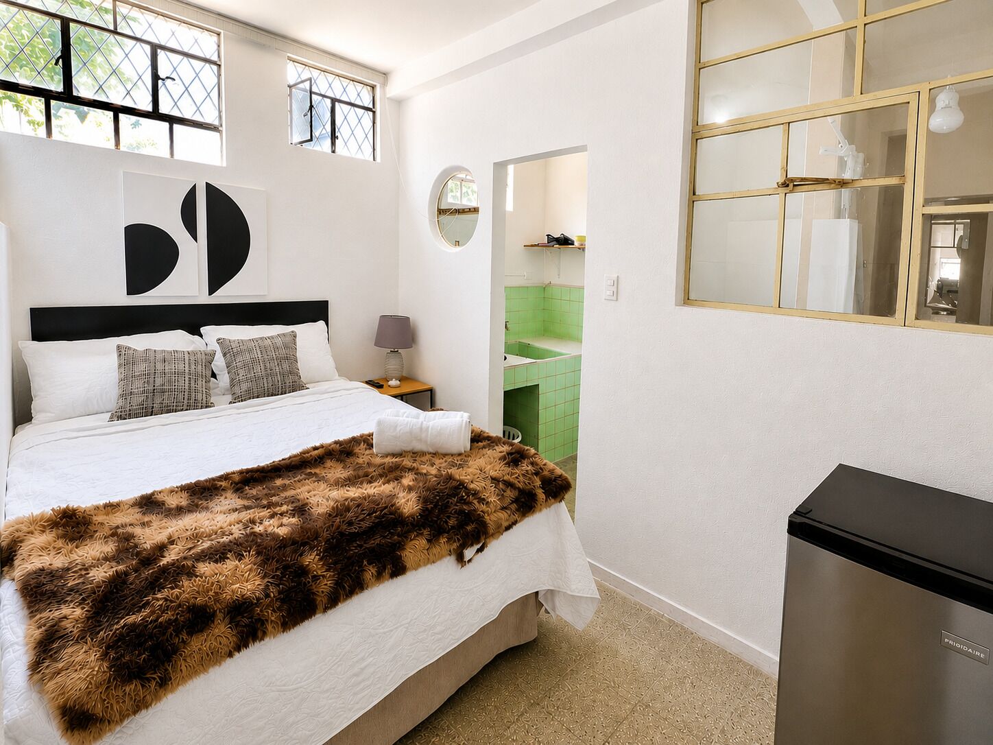 Un dormitorio con cama, mesita de noche, lámpara, espejo y refrigerador.