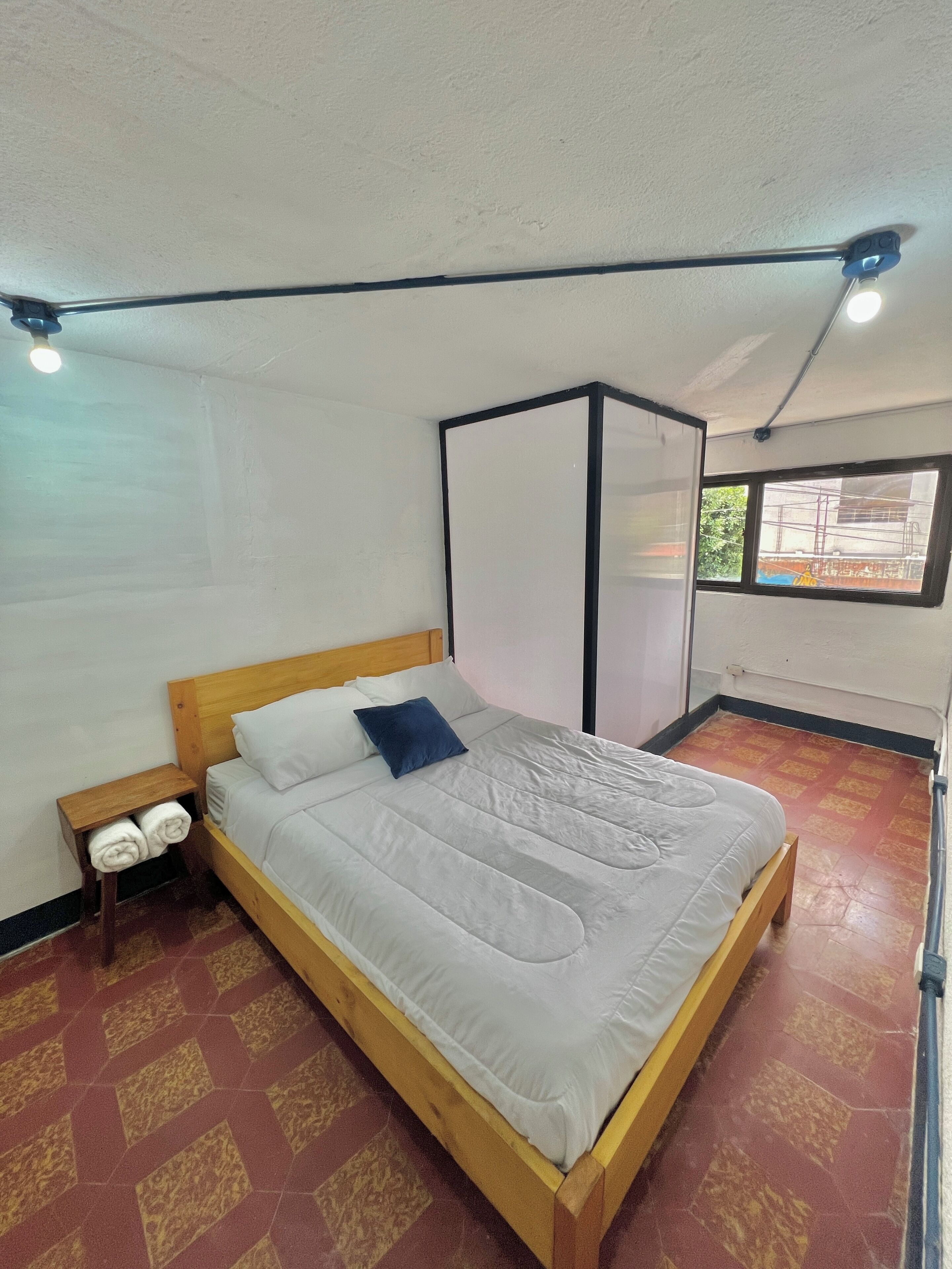 Un lit simple avec un cadre en bois et du linge de lit blanc, une table de chevet avec des serviettes, un ventilateur de plafond, une fenêtre donnant sur des immeubles et un sol carrelé.