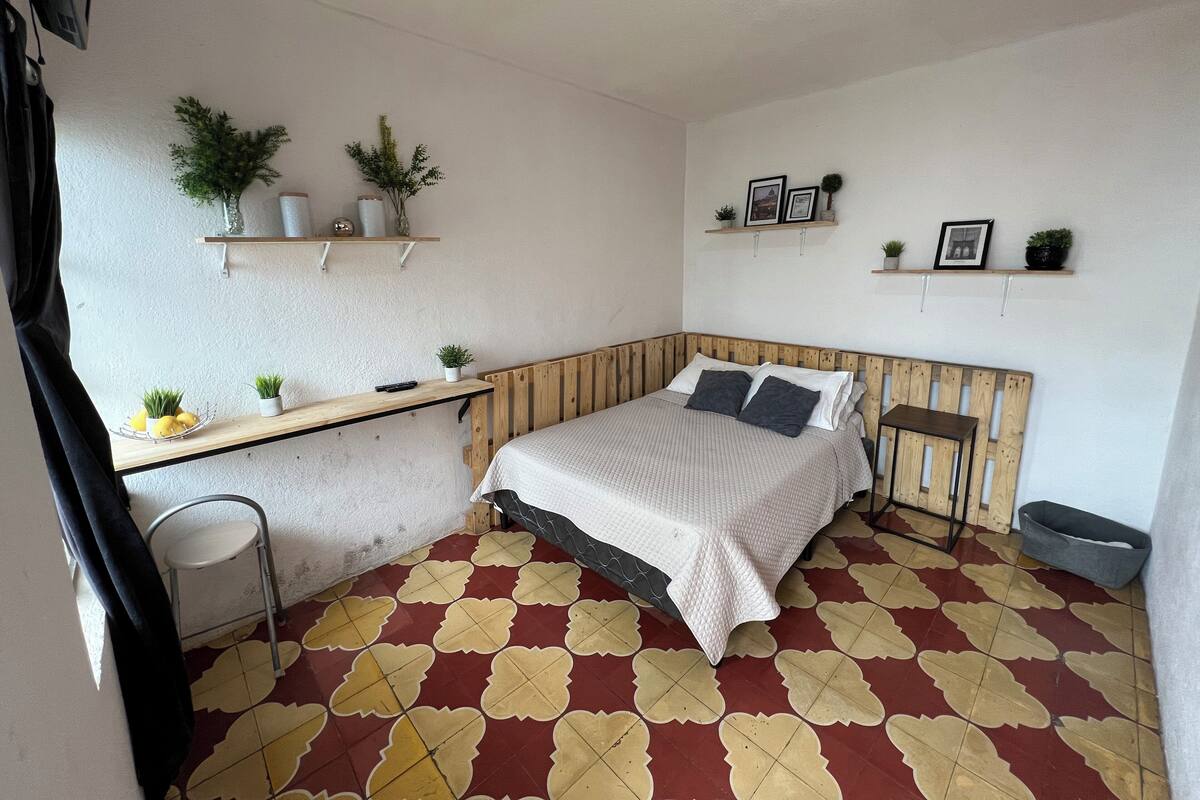 A small, neatly arranged bedroom with a bed, bedside table, and shelves with plants and pictures.