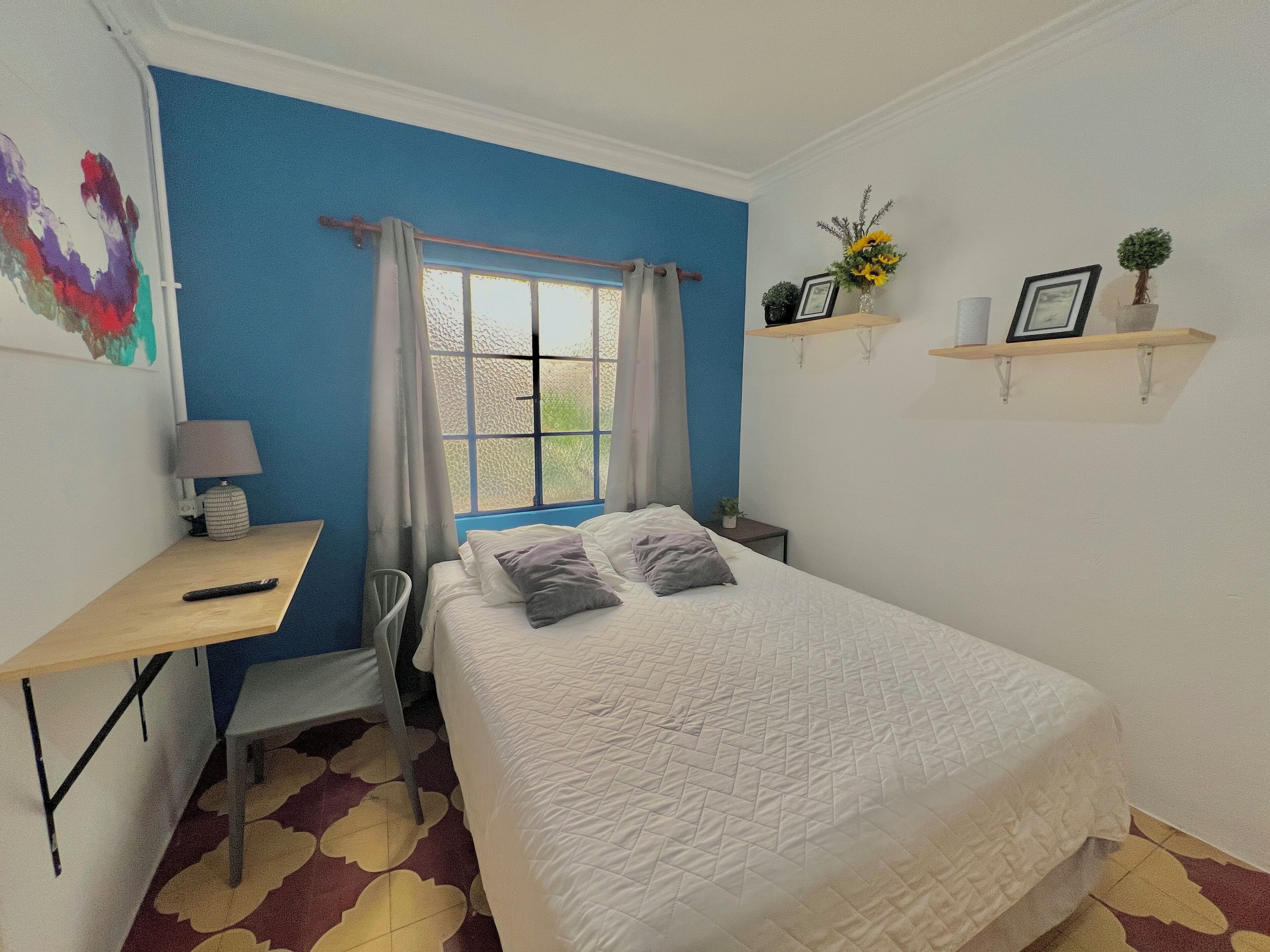 A bedroom with a bed, a desk, a chair, a window with curtains, and wall-mounted shelves with decorative items.