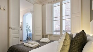 A neatly made bed with white linens and a brown bedspread, a window with white shutters, and a door leading to another room.