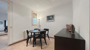 A dining area with a round table and four chairs, a window with curtains, a framed picture on the wall, and a small table with decorative items. - Stay U-nique Apartments Calabria (Barcelona)