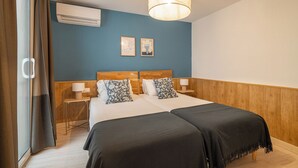 Cama doble con dos almohadas, mesita de noche con lámpara, aire acondicionado montado en la pared y dos cuadros enmarcados.