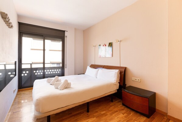 A neatly made bed with white linens and towels, a wooden nightstand with a drawer, and a window with blinds.