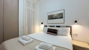 A neatly made bed with white linens and two towels, a bedside table with a lamp, and a framed picture on the wall.