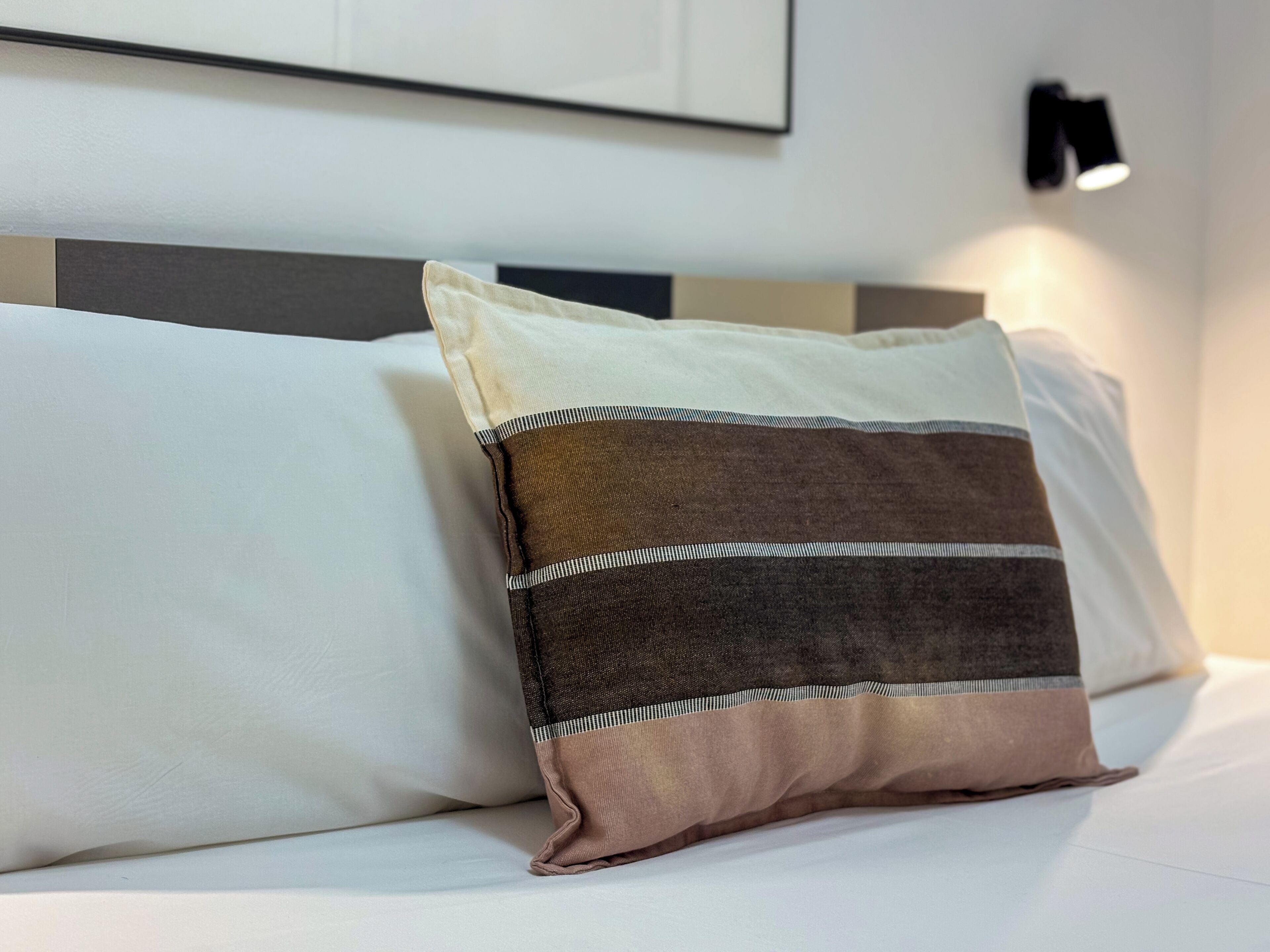 A bed with two pillows, one plain and one striped, in a room with a modern headboard and a wall-mounted light fixture.
