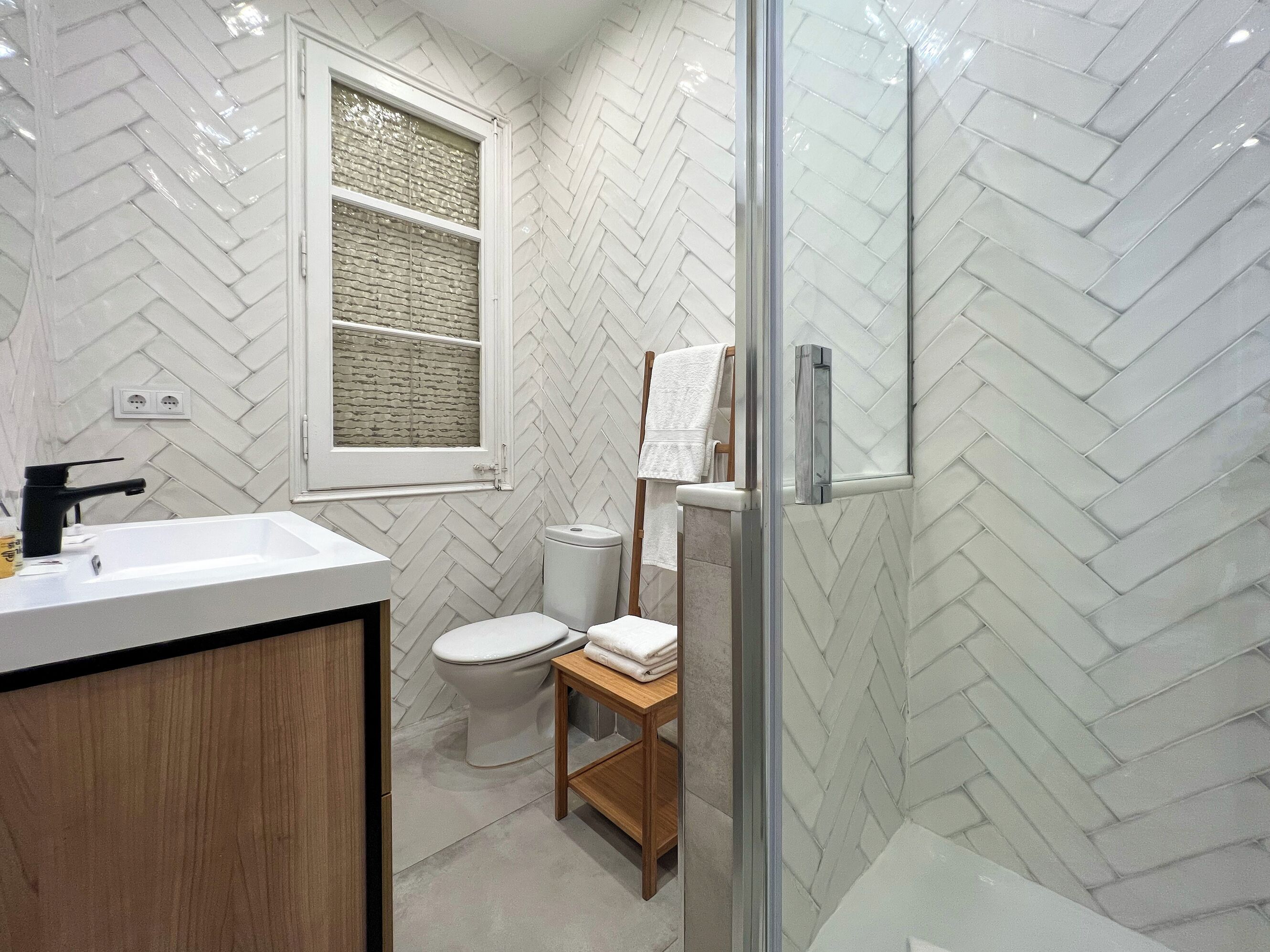 A modern bathroom with a herringbone tile pattern, a white sink, a toilet, and a glass shower enclosure.