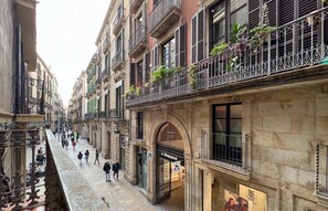 A narrow street with buildings on either side, featuring balconies with plants and wrought iron railings.