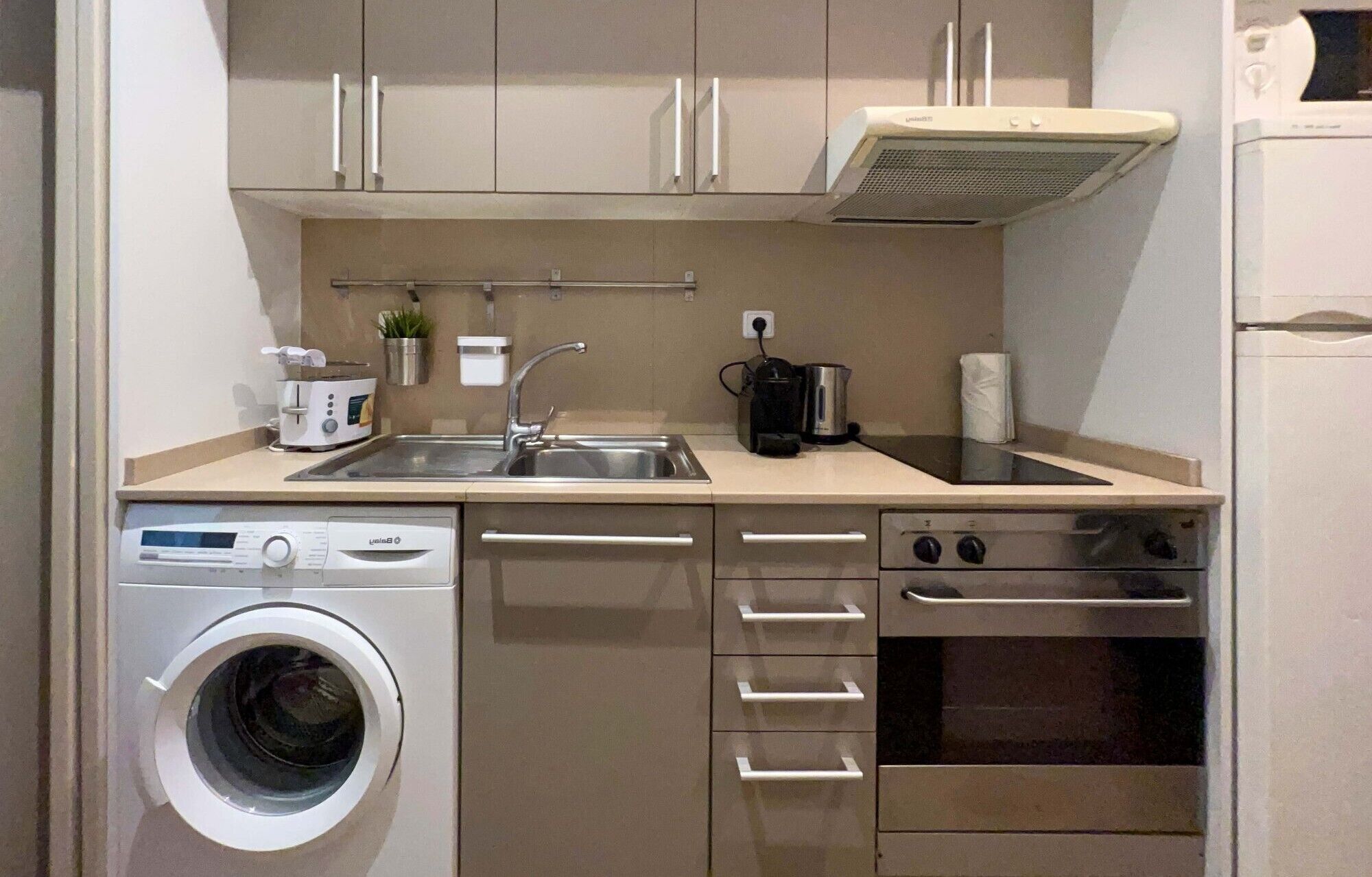 A compact kitchen with a washing machine, a coffee maker, a kettle, and a stove.