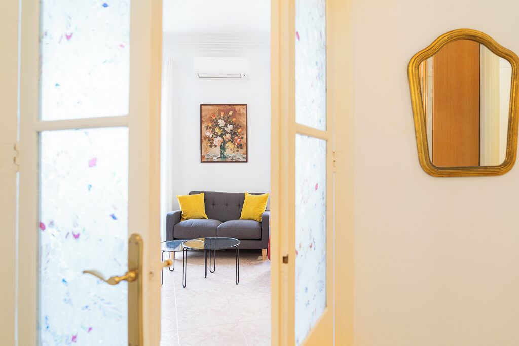 Une porte ouverte donne sur un salon où se trouvent un canapé gris, deux coussins jaunes, une petite table ronde et un tableau encadré accroché au mur.