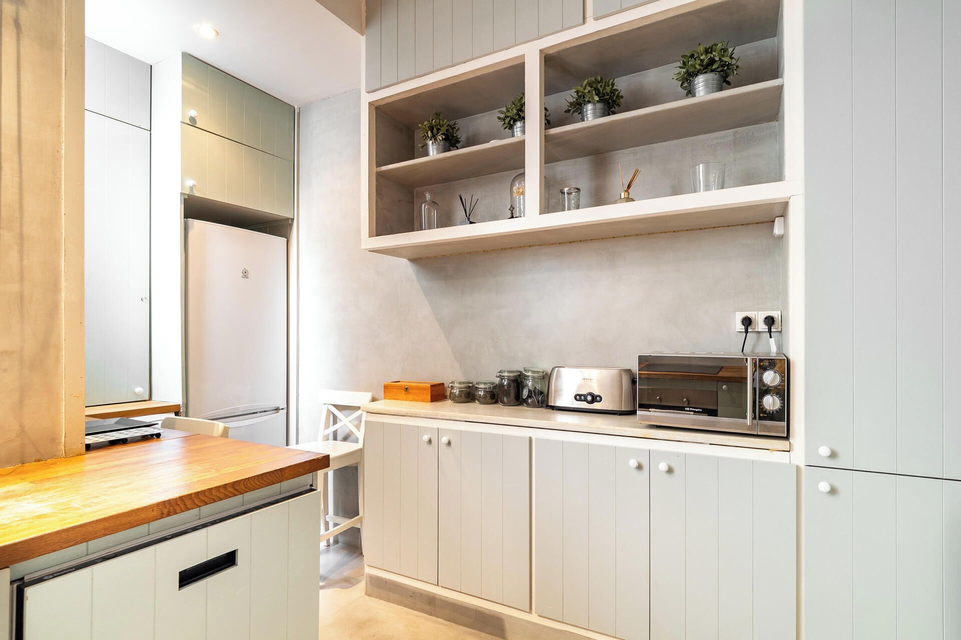 A modern kitchen with white cabinets, a wooden countertop, a toaster, a coffee maker, and potted plants on shelves.
