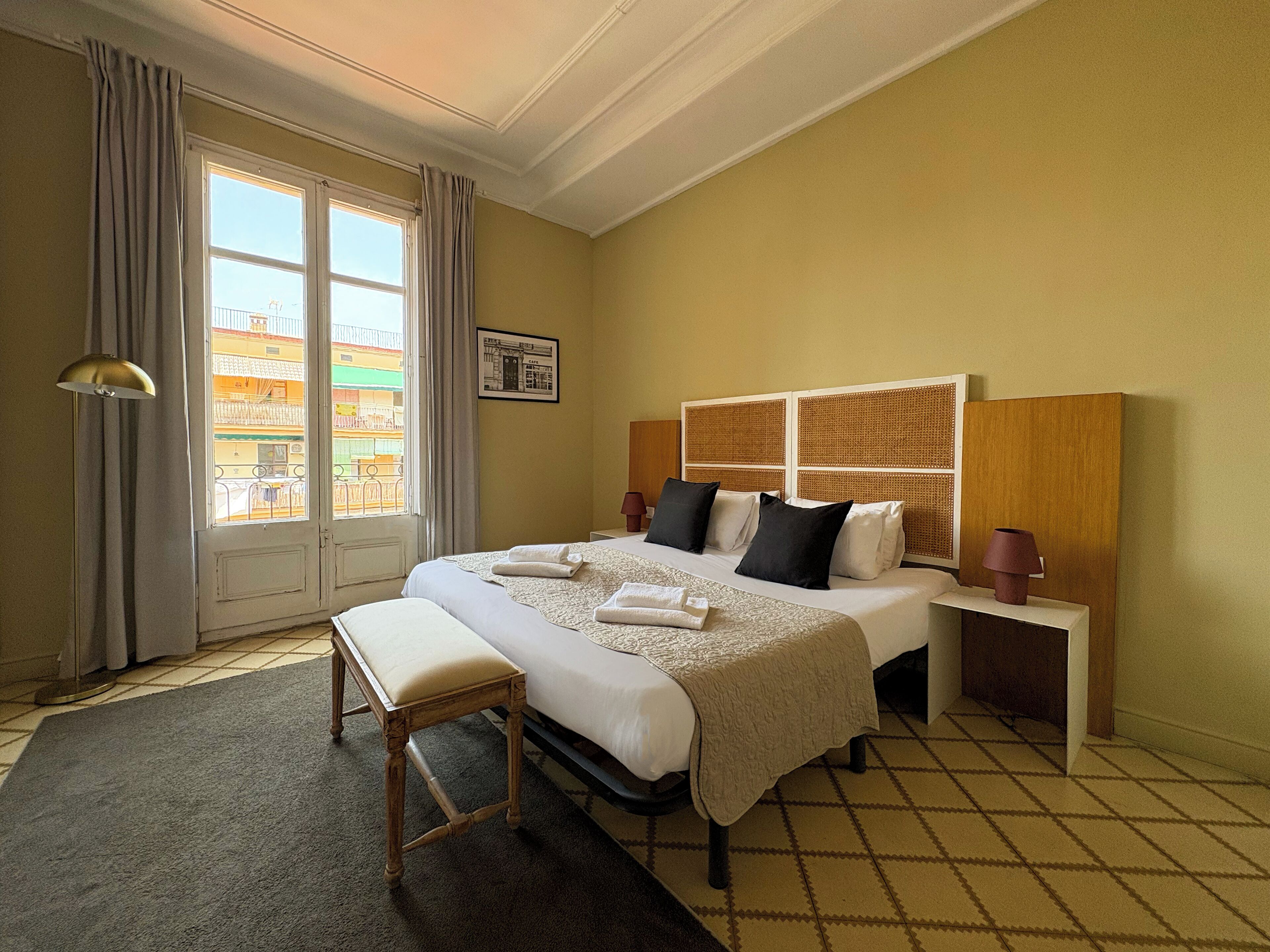 A hotel room with a large bed, a bedside table, a bench, and a window with curtains.