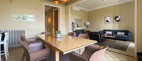 Un comedor con una mesa larga, varias sillas, lámparas colgantes, un radiador y un sofá al fondo.