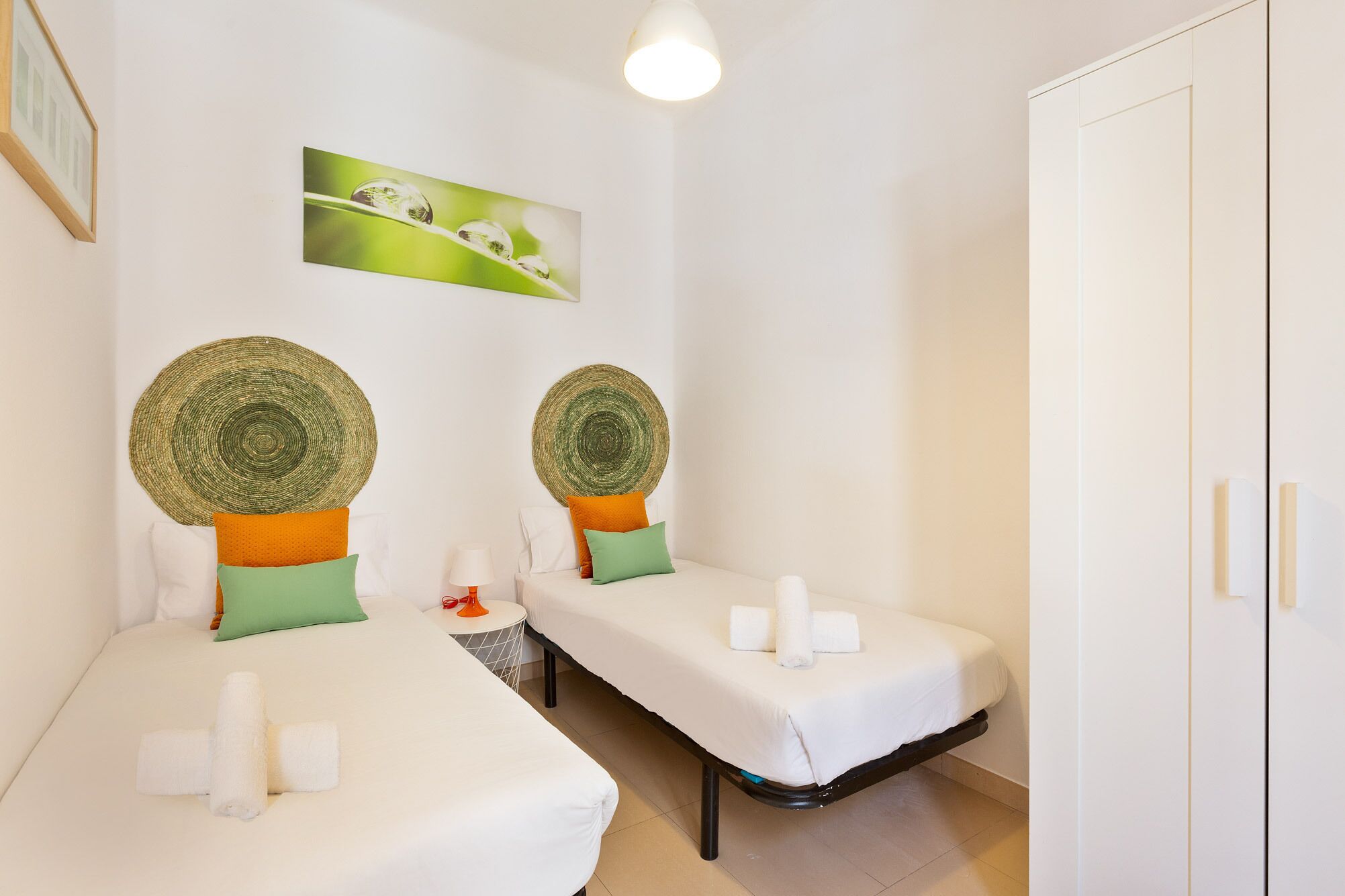 A hotel room with two single beds, each with a white mattress and a rolled towel. There are two woven wall decorations above the beds, a small round table with a lamp, and a framed picture on the wall.