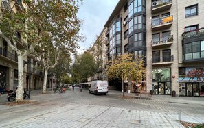 Una calle con una camioneta blanca estacionada en la acera, árboles a lo largo de la calle y edificios con balcones y escaparates.