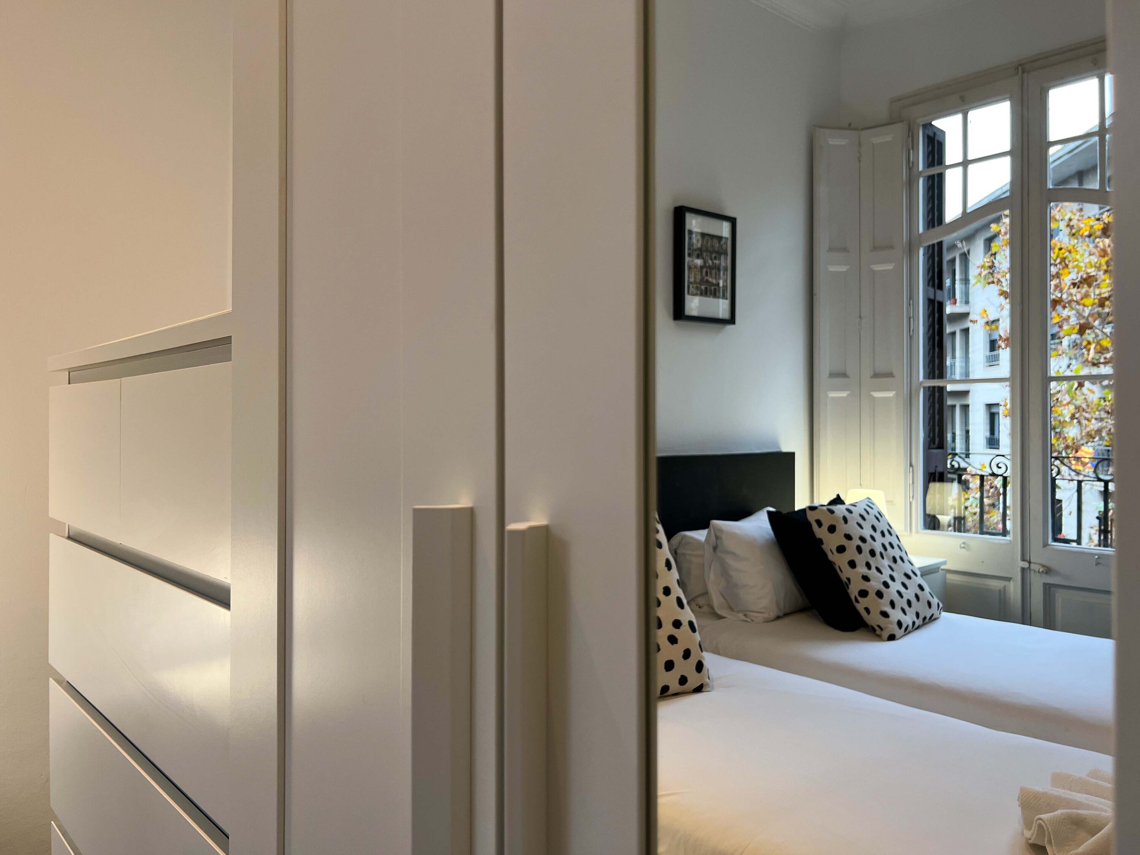 A bedroom with a bed, a nightstand, and a window with a view of buildings and autumn leaves.