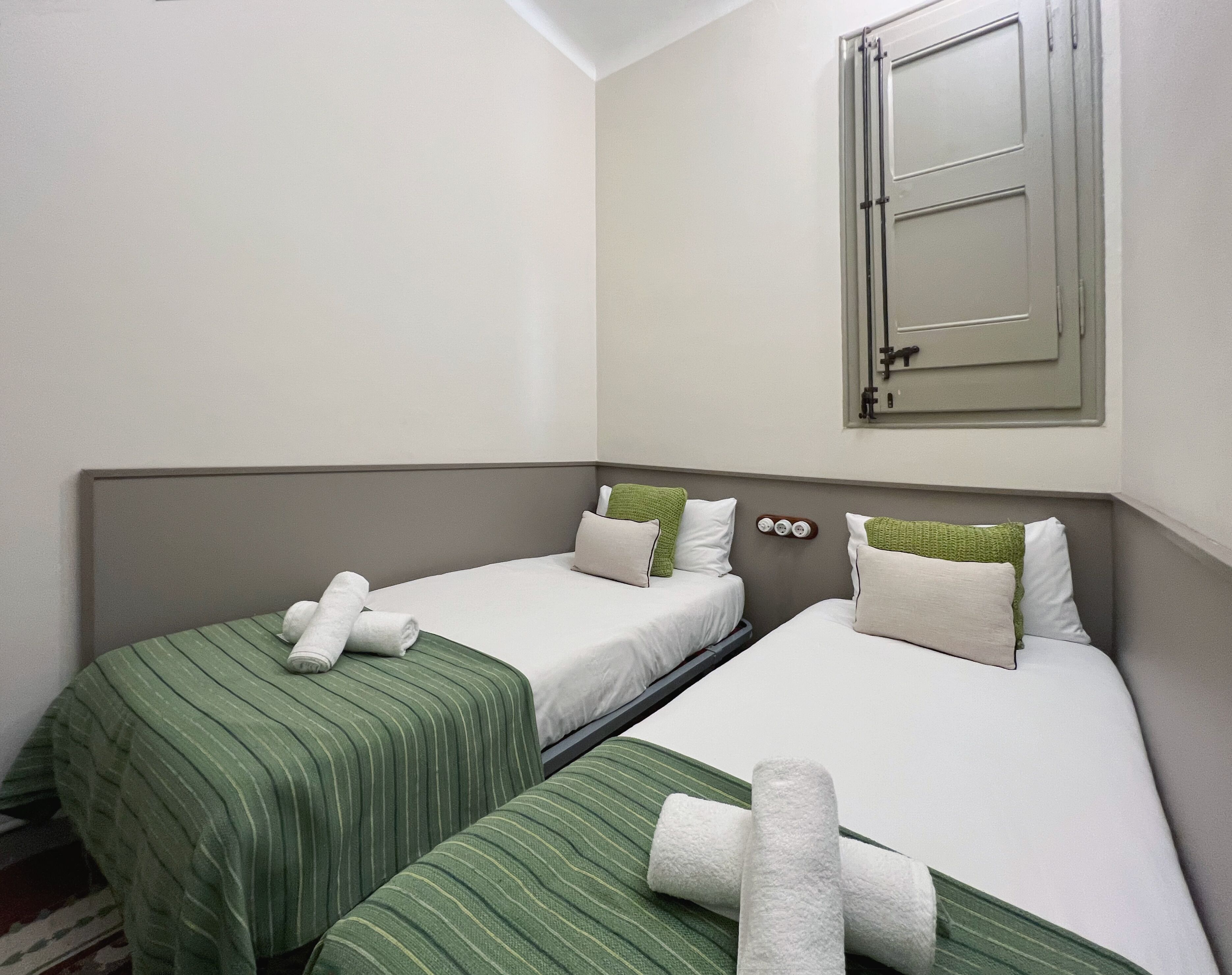 Una habitación de hotel con dos camas individuales, cada una con una colcha verde a rayas y ropa de cama blanca. Se ve una ventana con marco de madera e interruptor de luz en la pared.