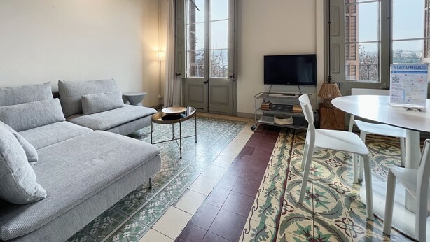 A living room with a grey sofa, a small round table, a TV, and a white table with chairs. - Stay U-nique Apartments Borbó II (Barcelona)