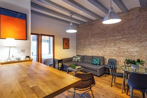 Una sala moderna con una mesa de comedor de madera, un sofá y una mesa de centro de vidrio. Hay dos lámparas colgantes, una pared de ladrillo y una puerta que da a otra habitación.