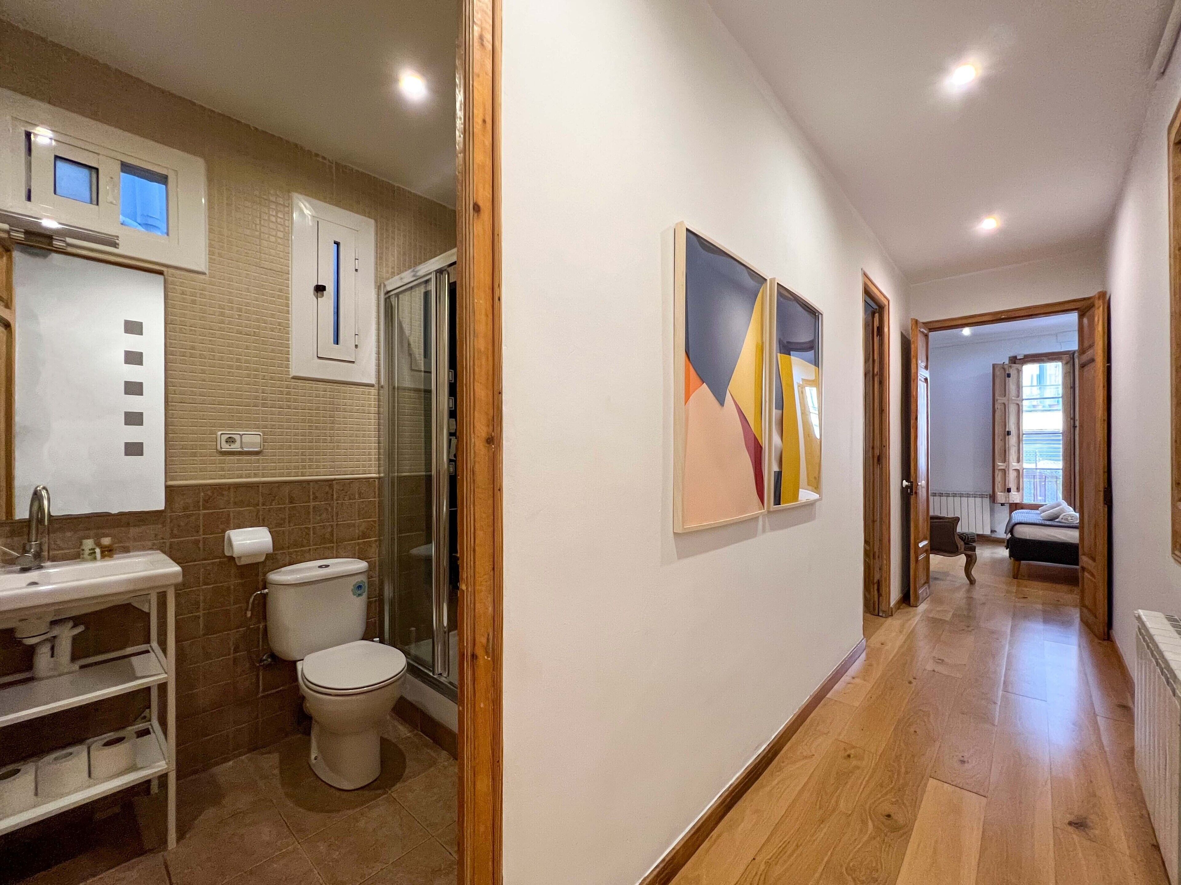 Een hal met aan de linkerkant een badkamer, voorzien van toilet, wastafel en douche. Er ligt een houten vloer, de muren zijn wit en het plafond heeft inbouwverlichting.