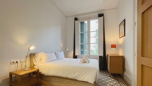 Quarto com cama, luminárias de cabeceira, mesa de cabeceira de madeira, uma mesinha, piso estampado e janela com cortinas.