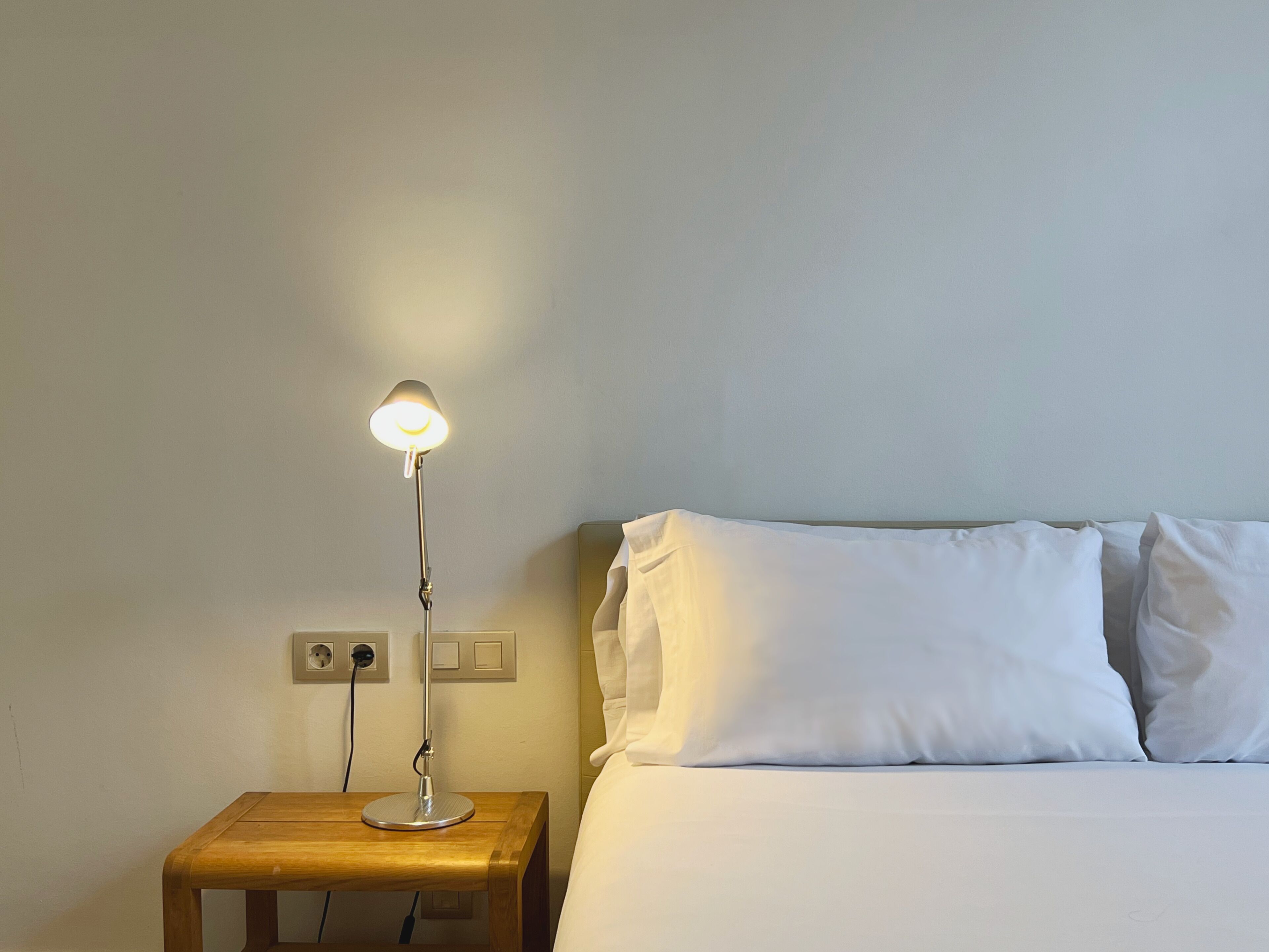Ein ordentlich bezogenes Bett mit weißen Leinen und Kissen, ein Nachttisch mit Lampe und eine Steckdose.