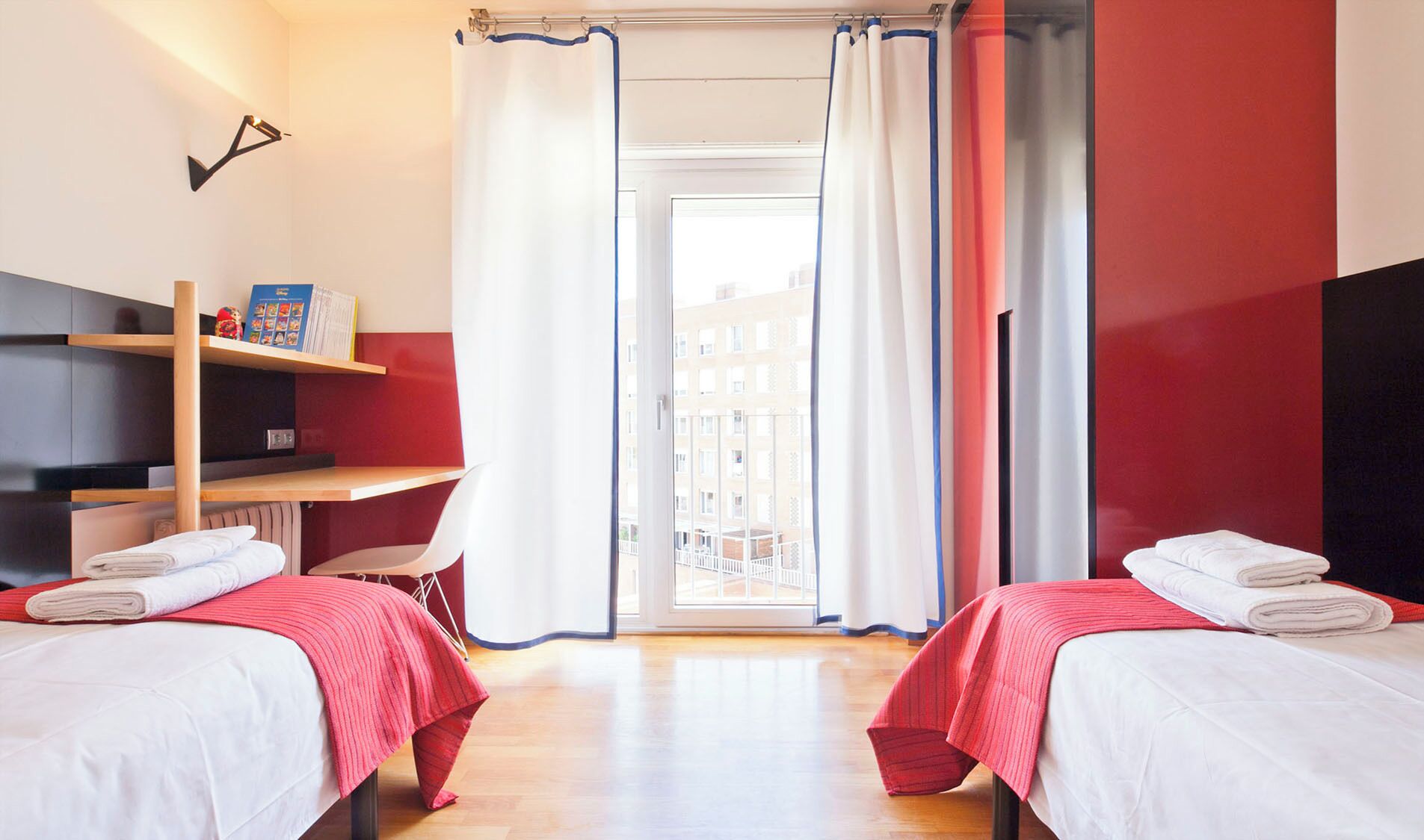 A hotel room with two beds, a bookshelf, a desk, and a view of buildings through a large window.