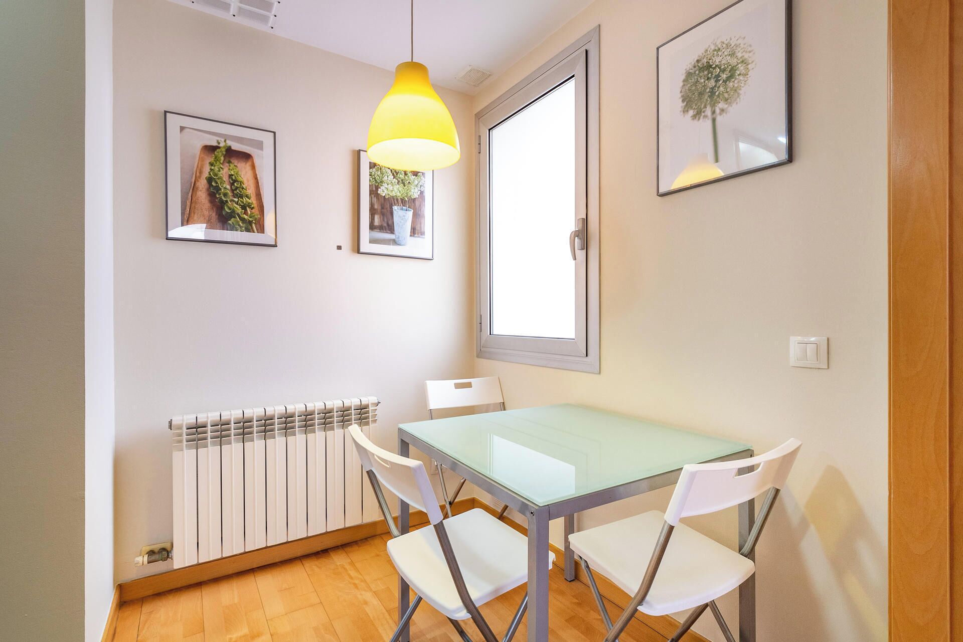 A dining area with a glass table and white chairs, a radiator, and two framed pictures on the wall.