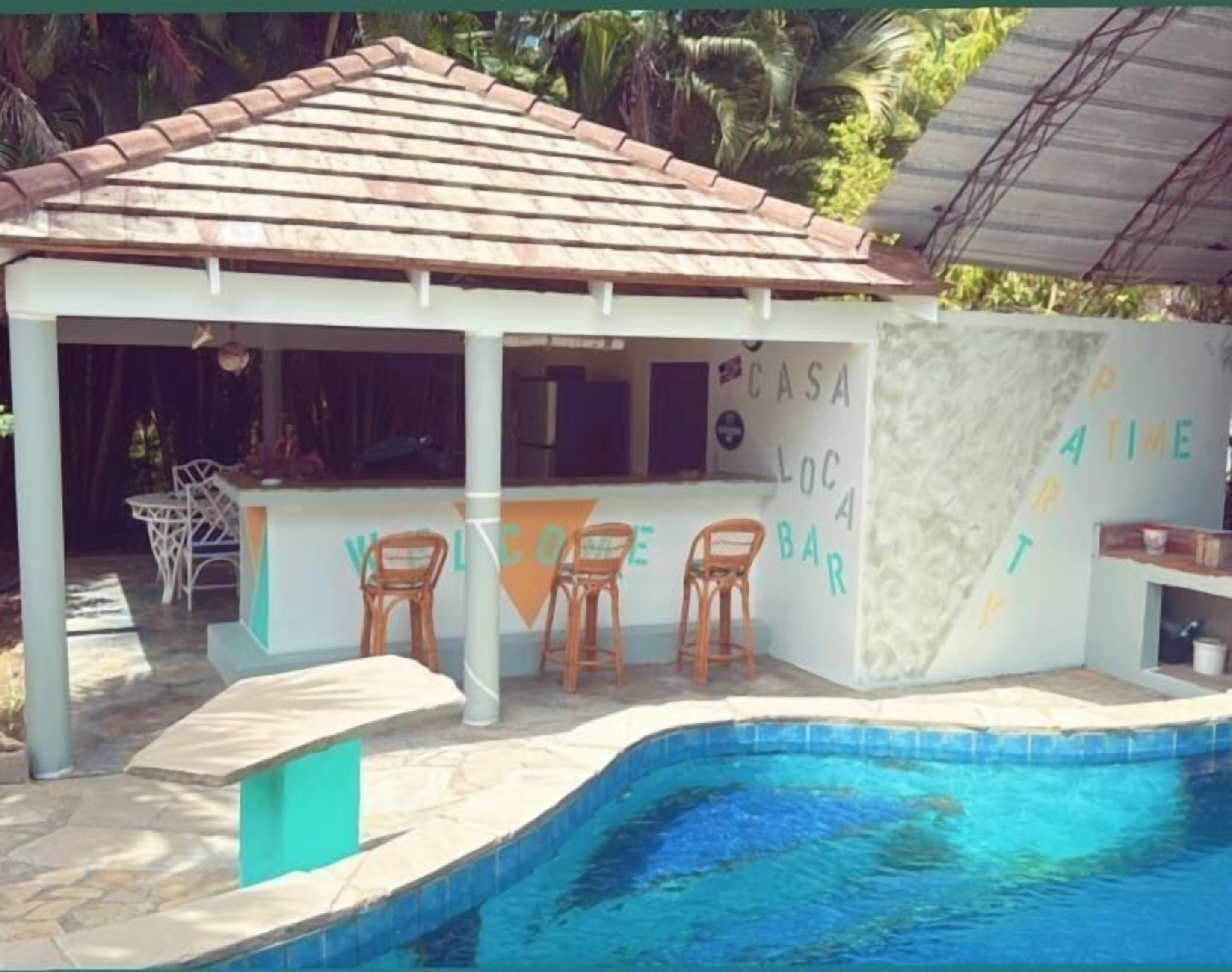 Un bar junto a la piscina con techo de azulejos, tres sillas de madera y un letrero que dice "CASA LOC BAR".