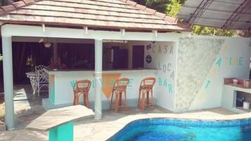 Un bar junto a la piscina con techo de azulejos, tres sillas de madera y un letrero que dice "CASA LOC BAR".