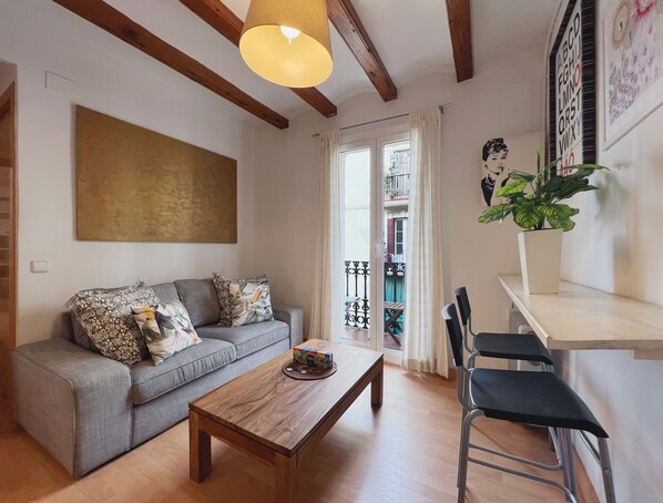 A living room with a sofa, a wooden coffee table, and a chair. There is a window with curtains, a plant, and a wall with a painting. - Stay U-nique Apartments Murillo (Barcelona)