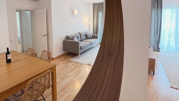 Een moderne woonkamer met een grijze bank, een houten eettafel met stoelen, en een grote spiegel die de kamer weerspiegelt.
