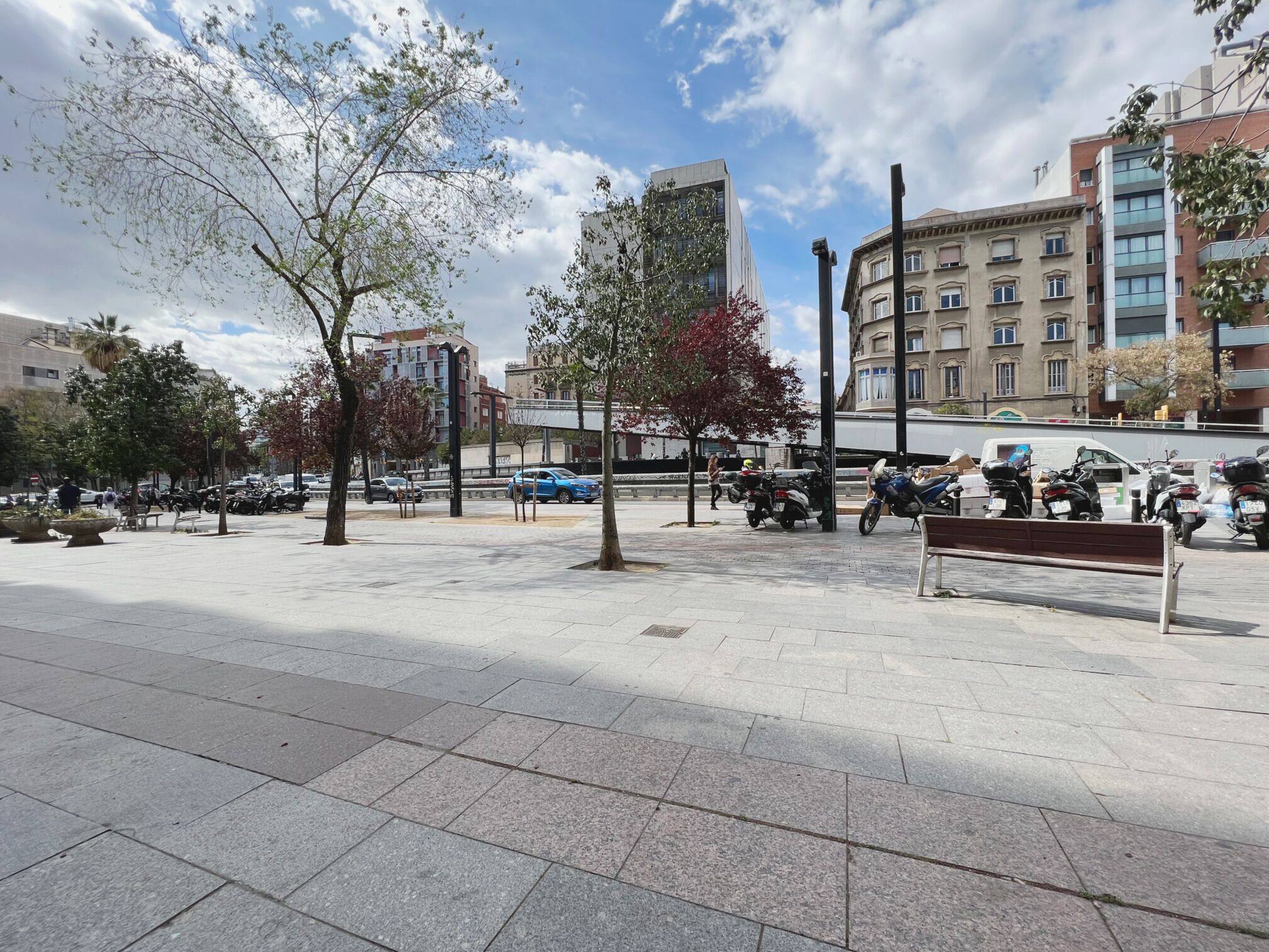 Une place urbaine dallée, agrémentée d’arbres, de bancs et de motos garées.