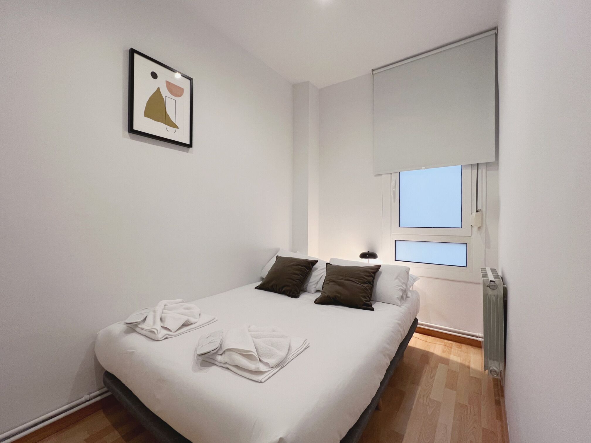 A neatly made bed with white linens and brown pillows, a framed abstract artwork on the wall, and a window with blinds partially open.