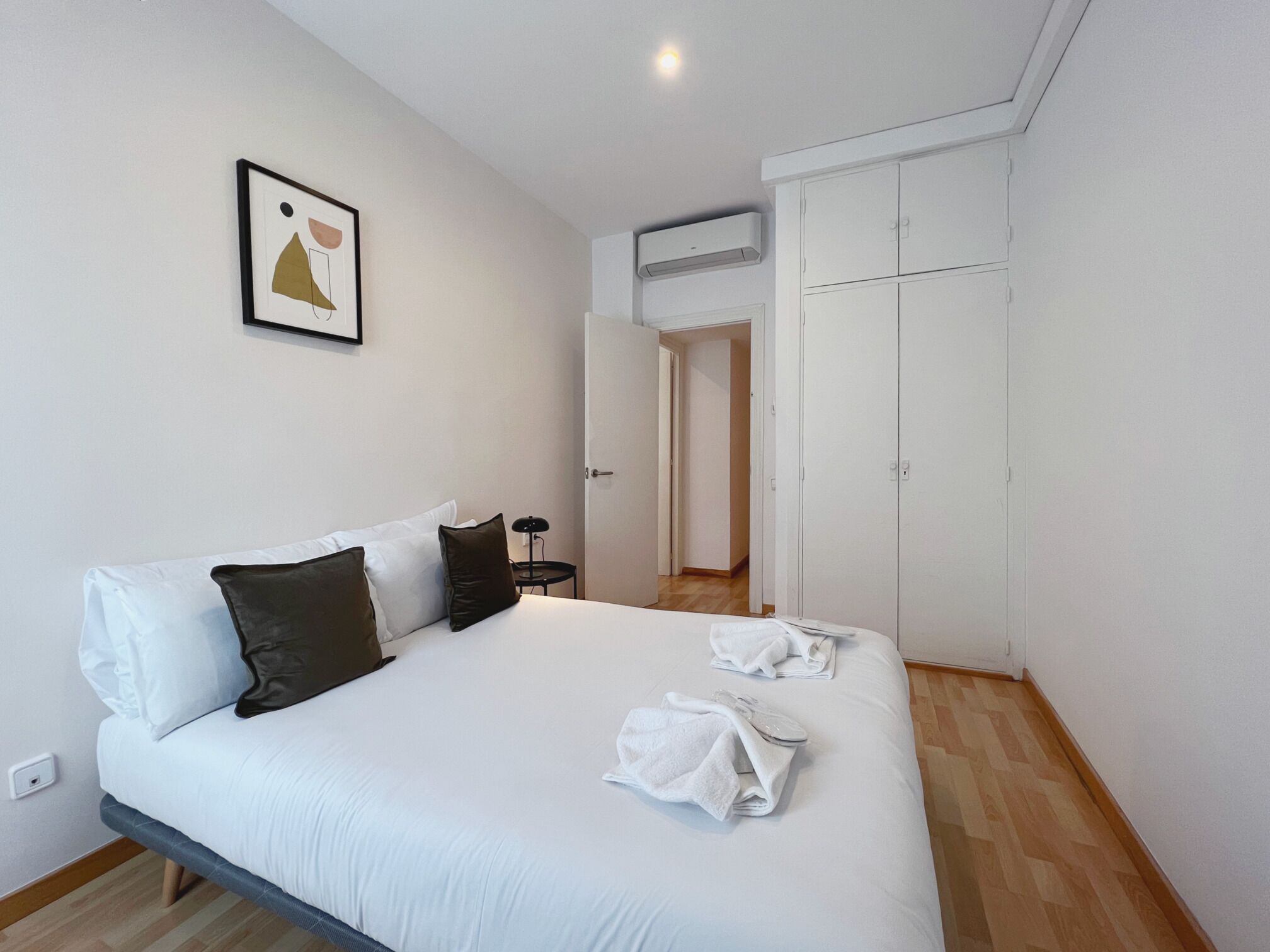 A neatly made bed with white and brown pillows, a framed picture on the wall, and a white wardrobe with a door leading to another room.