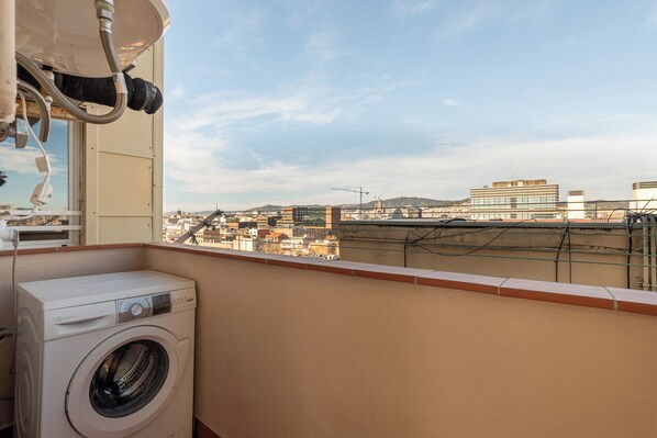 Un balcon équipé d’une machine à laver et d’un tuyau d’arrosage, donnant sur un paysage urbain avec des immeubles et des grues de construction.
