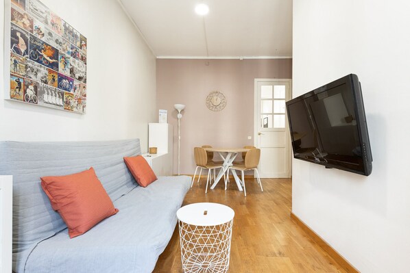 Ein modernes Wohnzimmer mit einem grauen Sofa, roten Kissen, einem runden weißen Beistelltisch, einem Holzesstisch mit vier Stühlen und einem an der Wand montierten Fernseher.