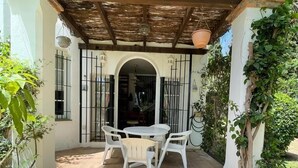 Eine überdachte Terrasse mit Tisch und Stühlen, ein Reetdach und Blick ins Haus durch einen Bogen.