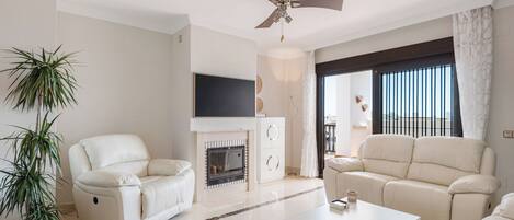 Una sala de estar moderna con chimenea, dos sofás de cuero blanco, una mesa de centro blanca y un ventilador de techo.