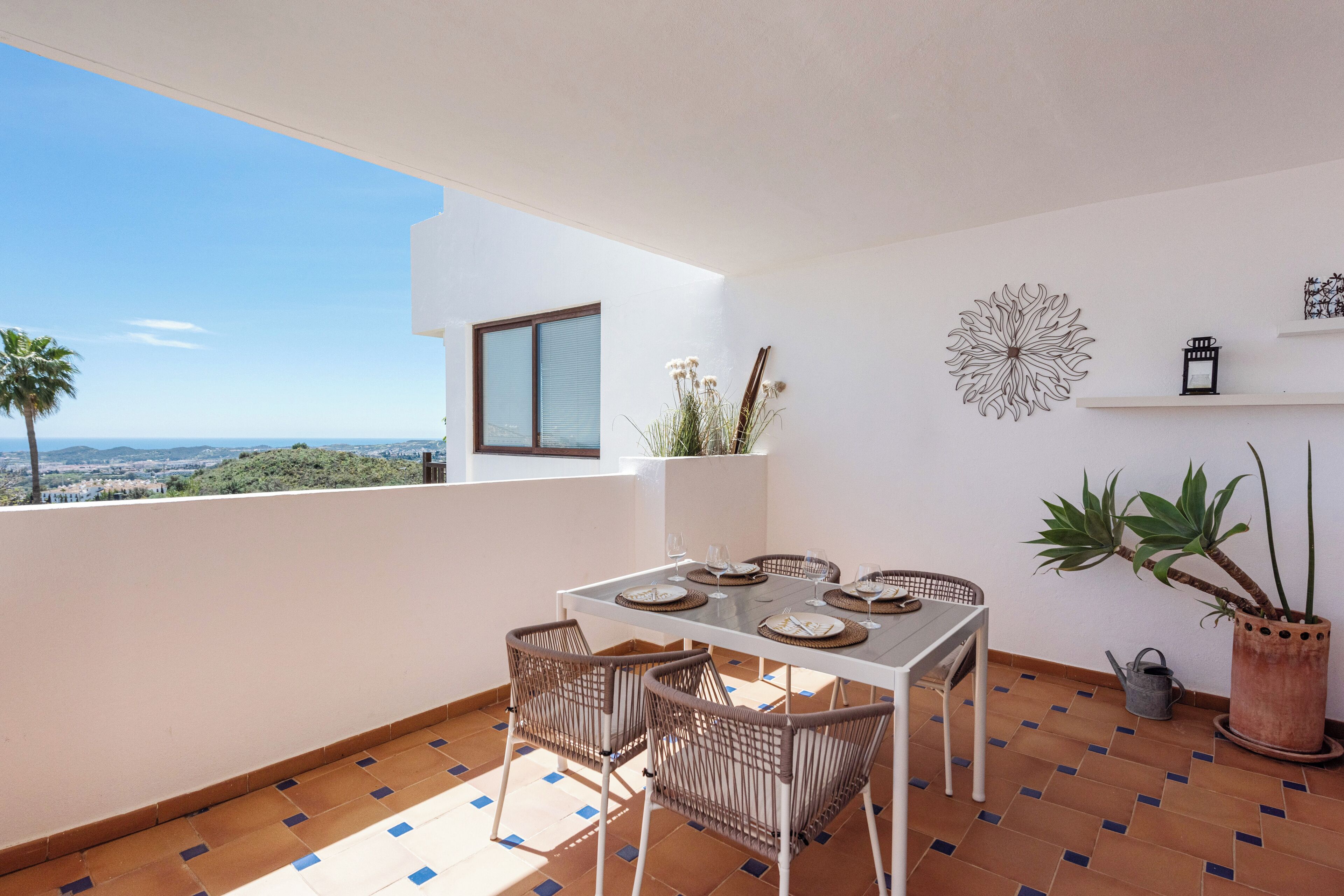 A balcony with a table set for four, surrounded by wicker chairs, a potted plant, and decorative wall art.